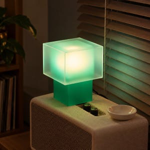 Może przedstawiać: Kwadratowa lampa stołowa z kloszem w kształcie sześcianu z matowego zielonego szkła na solidnej zielonej podstawie. Lampa emituje ciepłe, miękkie światło od wewnątrz, oświetlając półprzezroczysty zielony materiał. Stoi na prostokątnym głośniku o teksturze.
