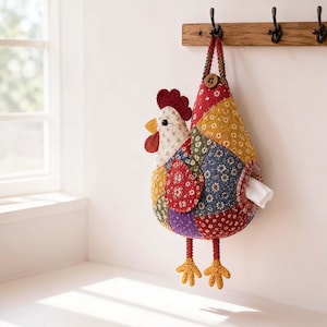 Puede incluir: Un dispensador de pared de tela con forma de gallina, hecho de varios parches acolchados con patrones florales en rojo, amarillo, azul y morado. La gallina tiene una cresta roja, un pico amarillo y dos patas amarillas de ganchillo colgantes. Un pañuelo blanco es visible a través de una abertura circular en el lateral, y el artículo está suspendido por una correa trenzada con un botón de madera.