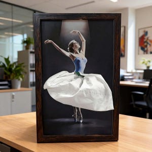 May include: A framed artwork featuring a ballerina in a blue leotard and a three-dimensional white paper skirt. The dancer is positioned under a spotlight against a dark background, captured in a dynamic pose with arms raised and pointed toes.