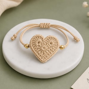 May include: A beige crochet heart bracelet featuring a central heart-shaped motif made of woven thread. The bracelet includes two small gold-toned metallic beads on either side of the heart and an adjustable sliding knot closure made of matching cord.