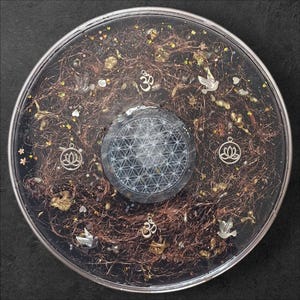Peut inclure: Un disque circulaire en résine présentant un motif central de fleur de vie entouré de fil de cuivre, de flocons dorés et de petites breloques argentées, dont des symboles Om, des lotus et des colombes incrustés dans une matière sombre et brillante.