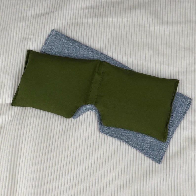May include: An olive green eye mask rests on top of a light blue sleep mask. The eye mask is rectangular with a curved cutout for the nose. The sleep masks are on a white, ribbed fabric.