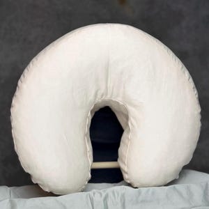May include: A crescent-shaped, off-white pillow with a smooth, solid-colored fabric cover. The pillow features a central U-shaped opening and a rounded, padded design, providing a consistent, neutral appearance throughout the entire surface.