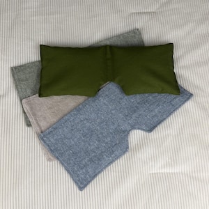 May include: An overhead shot shows four eye masks in shades of green, gray, and blue. The masks are made of soft fabric and are arranged on a white, ribbed surface. The masks are designed to block out light.