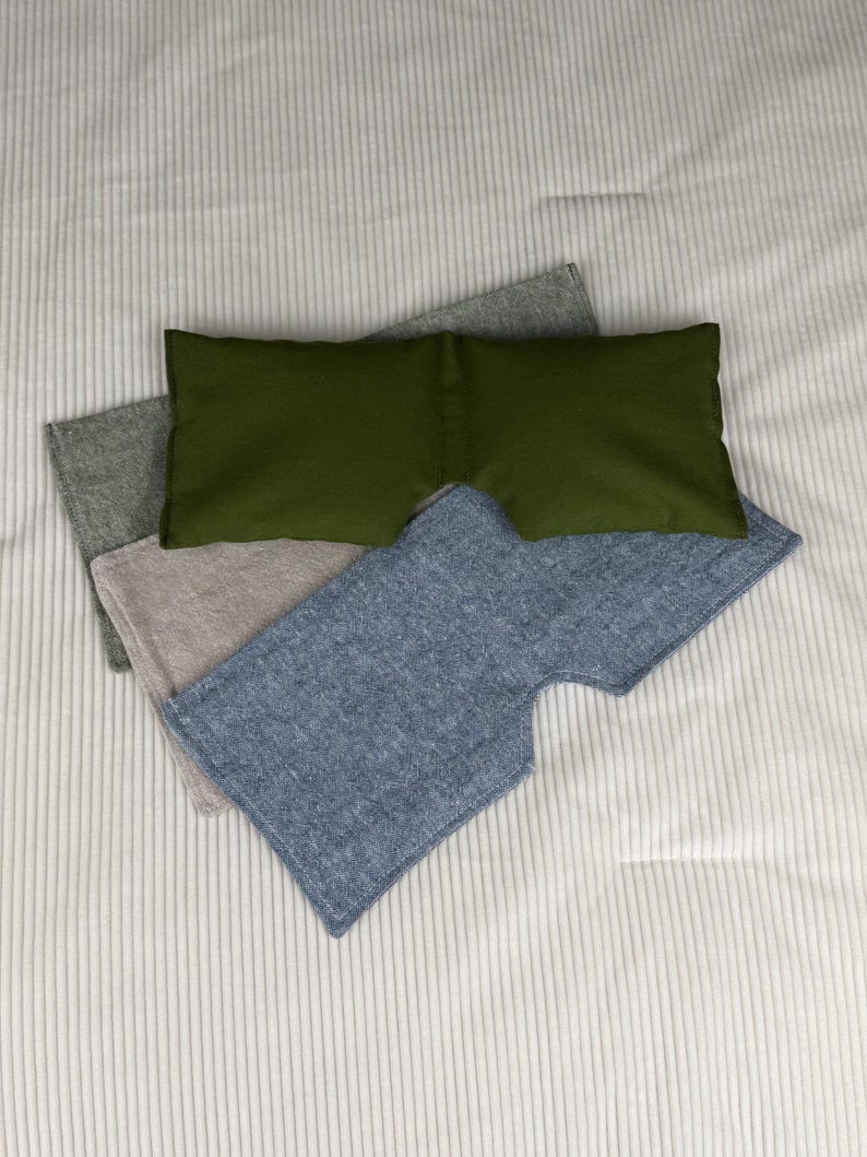 May include: An overhead shot shows four eye masks in shades of green, gray, and blue. The masks are made of soft fabric and are arranged on a white, ribbed surface. The masks are designed to block out light.