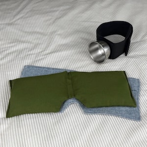 May include: An eye mask and a black cup holder with a silver cup are displayed on a striped surface. The eye mask is olive green and blue denim. The cup holder has a black strap with a silver cup attached.