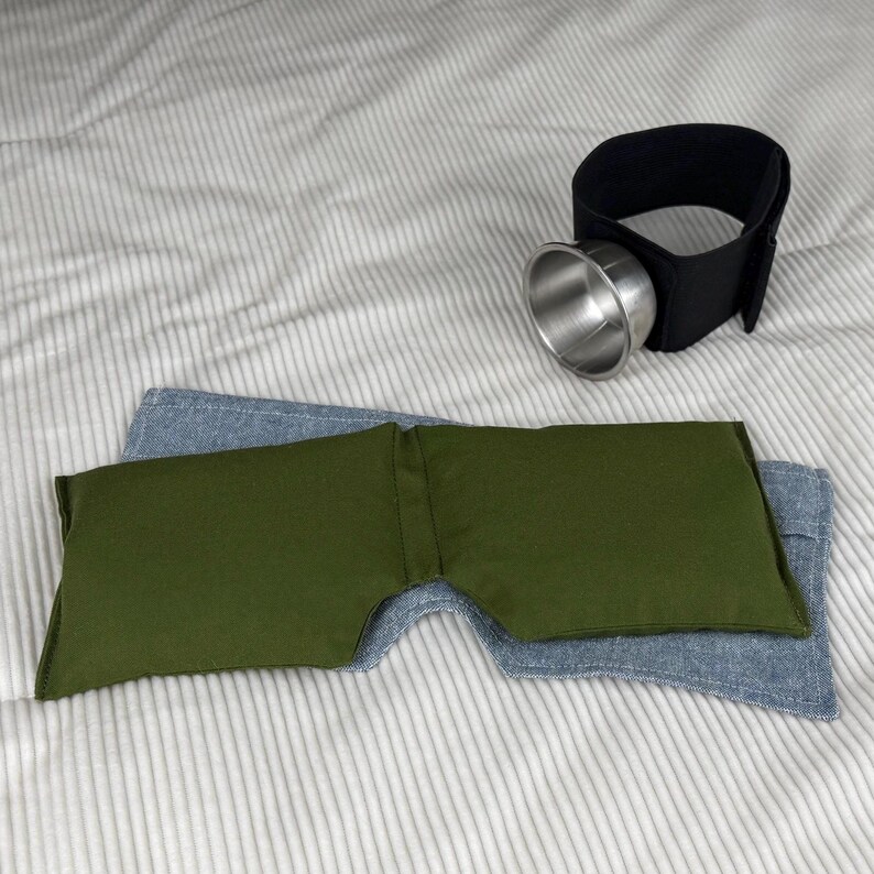 May include: An eye mask and a black cup holder with a silver cup are displayed on a striped surface. The eye mask is olive green and blue denim. The cup holder has a black strap with a silver cup attached.