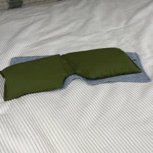 May include: An olive green eye pillow rests on a light blue fabric, both placed on a white and gray striped surface. The eye pillow is rectangular with a curved shape, designed to conform to the face. The fabric appears to be a soft, textured material.