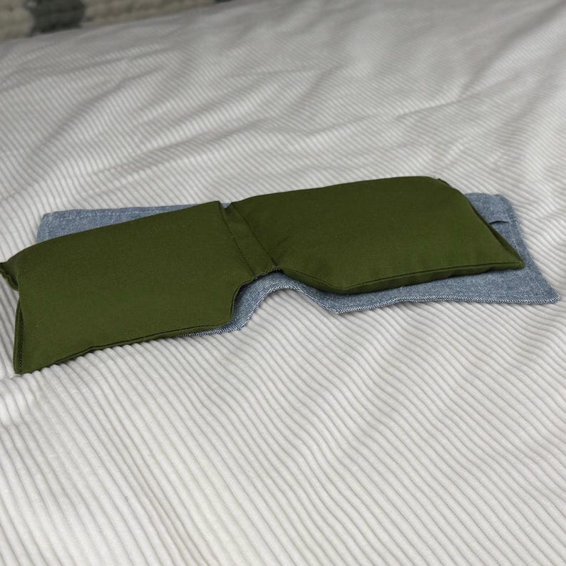 May include: An olive green eye pillow rests on a light blue fabric, both placed on a white and gray striped surface. The eye pillow is rectangular with a curved shape, designed to conform to the face. The fabric appears to be a soft, textured material.