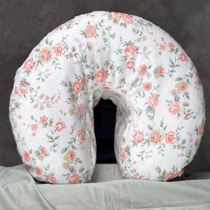 May include: A C-shaped nursing pillow featuring a white fabric base with a repeating floral pattern in shades of soft pink, peach, and muted green. The pillow has a curved, semi-circular design with a central opening, providing a cushioned support.