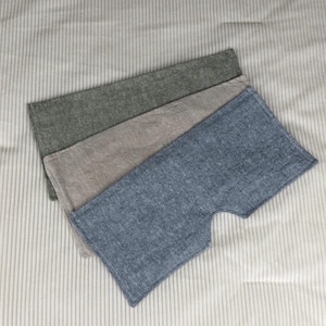 May include: Three rectangular, textured fabric pieces in shades of green, tan, and blue. The blue piece has a unique cutout shape. The fabrics are arranged on a light gray, vertically-striped surface.