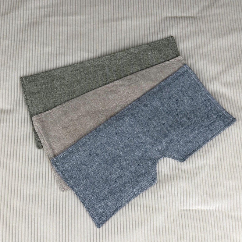 May include: Three rectangular, textured fabric pieces in shades of green, tan, and blue. The blue piece has a unique cutout shape. The fabrics are arranged on a light gray, vertically-striped surface.