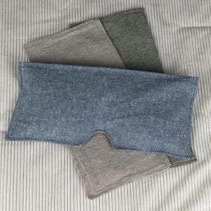 May include: A collection of eye masks in shades of blue, gray, and green, arranged on a striped fabric. The blue mask is in the foreground, with a cutout for the nose. The other masks are folded and layered behind it.