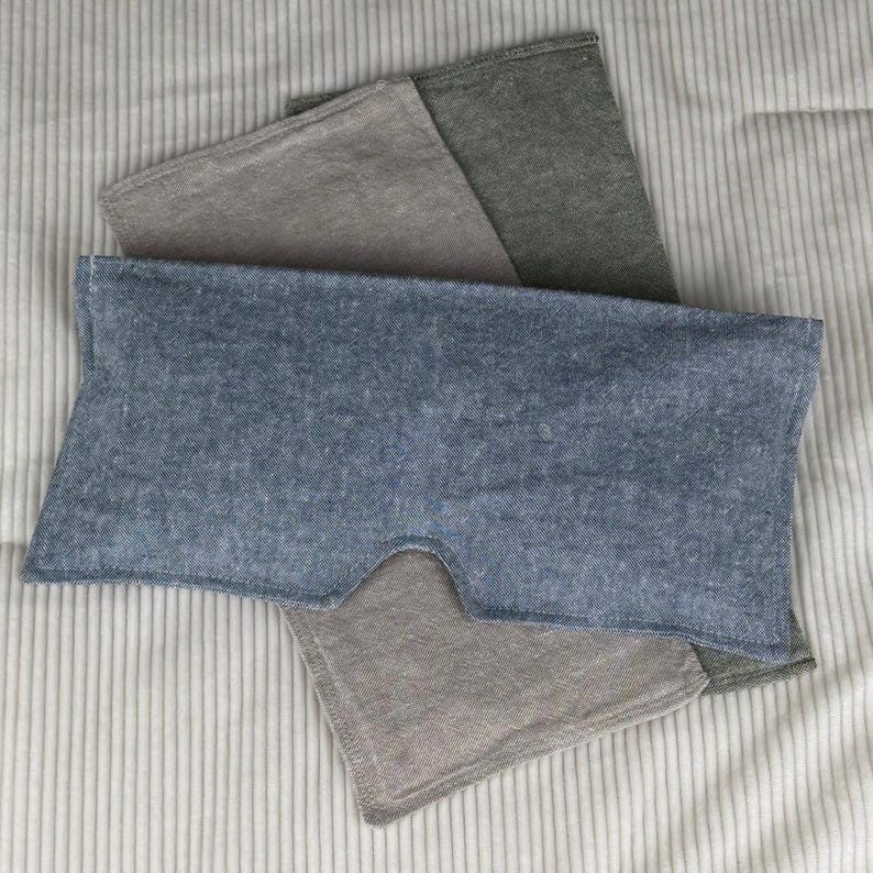 May include: A collection of eye masks in shades of blue, gray, and green, arranged on a striped fabric. The blue mask is in the foreground, with a cutout for the nose. The other masks are folded and layered behind it.