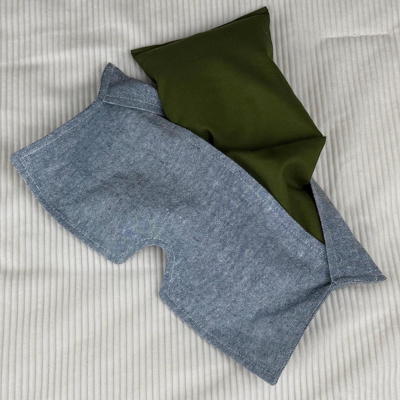May include: Two fabric eye masks, one blue and one olive green, rest on a white, ribbed surface. The blue mask has a rectangular shape with a curved cutout for the nose. The green mask is rectangular.