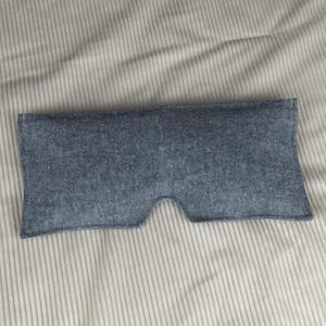 May include: A blue eye mask made of textured fabric. The mask has a curved shape to fit around the eyes and nose, with a cutout for the nose. The background is a light-colored, striped fabric.