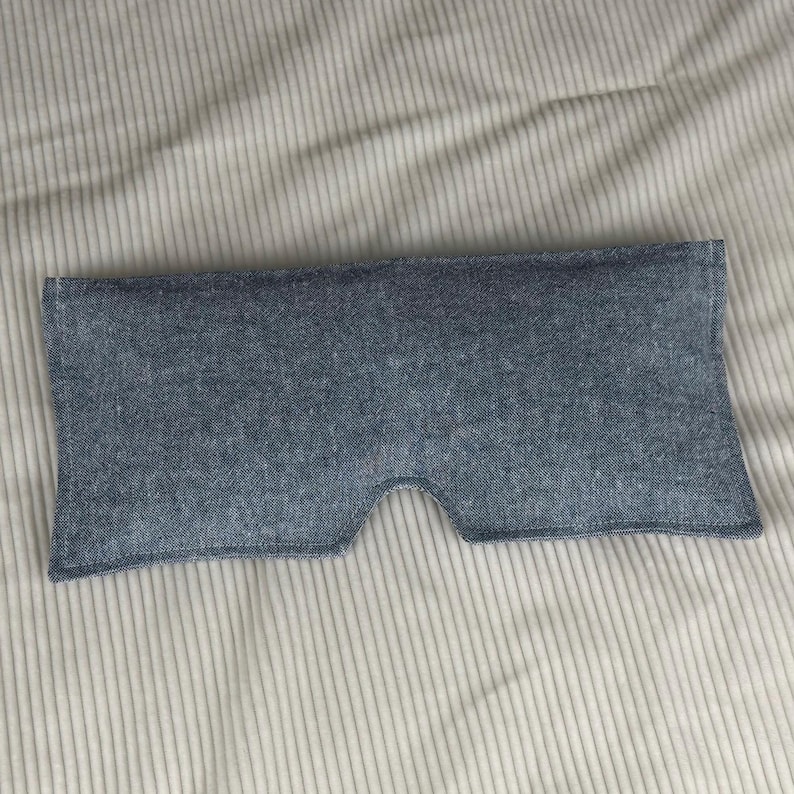 May include: A blue eye mask made of textured fabric. The mask has a curved shape to fit around the eyes and nose, with a cutout for the nose. The background is a light-colored, striped fabric.