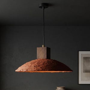 May include: A pendant light featuring a wide, shallow, textured terracotta-colored shade with a rough, uneven surface. The shade is attached to a dark brown wooden block connector, suspended by a black cord from a matching black ceiling canopy.