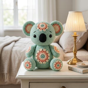 May include: A crocheted koala stuffed animal in light mint green yarn with a black nose and black eyes. The koala features decorative floral-patterned crochet patches in shades of cream and soft pink on its ears, chest, arms, and feet.