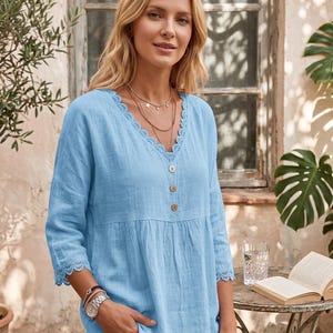 May include: A light blue blouse with a V-neckline and lace trim. The blouse has three-quarter sleeves and wooden buttons down the front. The model is wearing a watch and layered necklaces. The background includes a table with a glass of water and a book.