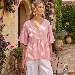 May include: A woman wearing a light pink V-neck top with ruffled sleeves and white linen pants. The top has a gathered waist. She is wearing gold earrings. The background features a stone building with purple flowers and potted plants.