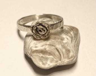 May include: A silver ring featuring a detailed rose design. The ring's band has a textured finish, and the rose is the focal point. The ring is displayed on a light-colored, textured surface, showcasing the jewelry's craftsmanship.