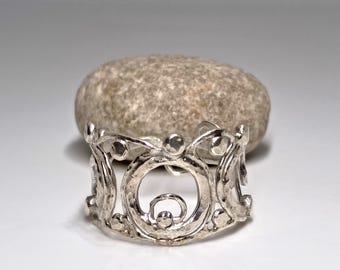 May include: A silver-colored ring with an openwork design featuring circular and swirling patterns. The ring is in focus, with a blurred, textured stone in the background. The ring's design includes small, raised circular details.