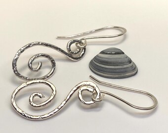 May include: A pair of silver swirl earrings with hammered texture, displayed with a small, gray and white striped seashell. The earrings have a hook closure. The jewelry is set against a white background.