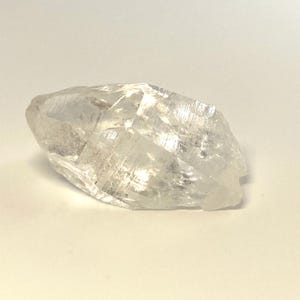 May include: A clear, crystalline quartz specimen with a rough, faceted surface. The quartz is translucent, allowing light to pass through, and has a natural, unpolished appearance. The overall shape is elongated and irregular, showcasing the raw beauty of the mineral.