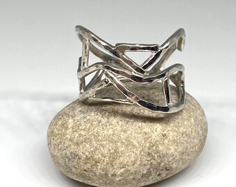 May include: A wide silver ring featuring an openwork design composed of interconnected, hammered metal bands. The surface of the silver has a textured, reflective finish with irregular, organic shapes forming a cage-like structure around the finger.