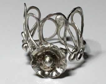 May include: A silver-colored ring with an openwork design. The ring features a floral element at the base and an intricate, looping pattern that forms the band. The metal has a slightly textured appearance, giving it a handcrafted feel.