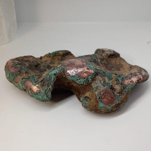 May include: A natural, irregularly shaped copper specimen with a mix of brown, green, and copper tones. The surface has a textured appearance, with areas of oxidation creating a unique, organic aesthetic. The piece is displayed on a white surface.