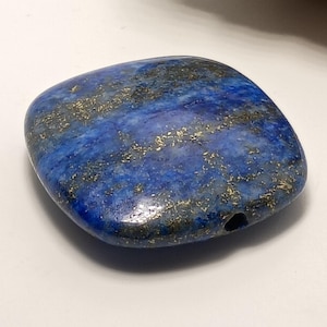 May include: A polished, square-shaped lapis lazuli gemstone. The stone is a deep blue color with gold flecks and veins throughout. A small hole is drilled through the stone, suggesting it is a bead or pendant.