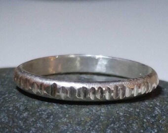 May include: A silver ring with a textured band. The ring has a hammered or etched design around the outer edge, creating a unique, handcrafted look. The ring is displayed on a dark gray surface.