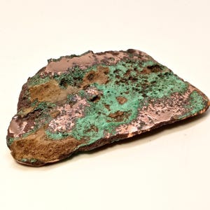 May include: A rough-hewn, irregularly shaped piece of copper with a mix of colors. The surface displays a blend of copper, brown, and vibrant green hues, suggesting oxidation. The edges are uneven, adding to its natural, raw appearance.