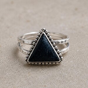 May include: A silver ring featuring a triangular black stone set in a beaded frame. The band has a split design with a hammered texture on the metal surface. The central triangle is dark, opaque, and smooth, contrasting with the metallic silver.