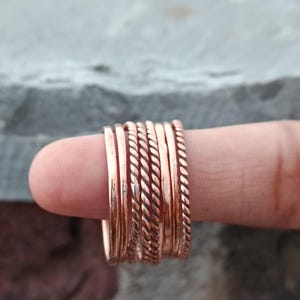 May include: A set of six individual copper rings worn together on a finger. The collection includes four smooth, polished bands and two twisted, rope-textured bands, all featuring a warm, metallic copper finish with a reflective surface.