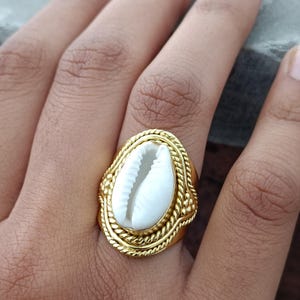 Peut inclure: Une bague dorée ornée d'un coquillage cauri blanc en son centre. Le coquillage est enchâssé dans un cadre ovale décoré d'une texture en corde torsadée et de petites perles sur le bord extérieur, formant une monture métallique détaillée.