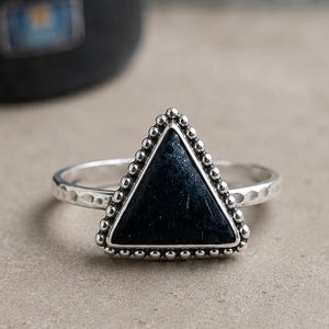 May include: A silver ring featuring a triangular black stone centrepiece. The stone is framed by a beaded silver border. The ring band has a hammered, textured finish, providing a distinct contrast to the smooth, dark triangular gemstone.