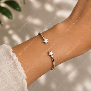 May include: A silver-colored cuff bracelet with star-shaped accents. The open-ended design features a star at each end, creating a celestial theme. The bracelet is worn on a wrist, showcasing its simple elegance.