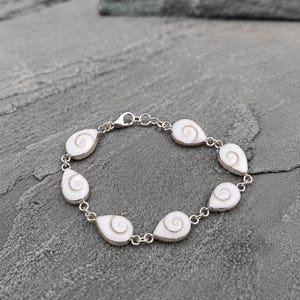 Puede incluir: Una pulsera de cadena de plata con siete dijes de concha de ojo de Shiva en forma de lágrima, de color blanco y tostado. Cada concha está engastada en un bisel de plata y conectada por pequeños eslabones circulares de plata.