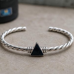 May include: A silver cuff bracelet featuring a twisted metal band design. The center of the bracelet is adorned with a black triangular stone inlay, flanked by two silver wire-wrapped accents on either side of the triangle.