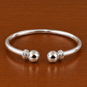 Puede incluir: Una pulsera abierta de tono plateado pulido con una banda circular lisa. Los dos extremos de la pulsera terminan en grandes remates esféricos, cada uno acentuado con tres sutiles anillos grabados en paralelo en la base de la esfera.
