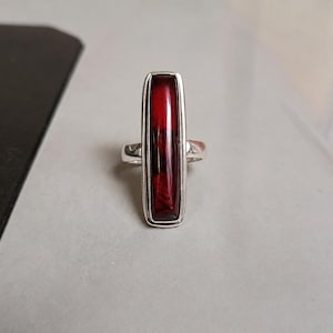 May include: A silver ring featuring a long, rectangular, deep red stone set vertically. The stone has a smooth, polished surface with dark, mottled inclusions visible within the red colour. The metal band is plain and polished, holding the stone.
