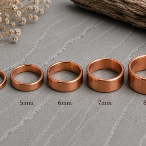 May include: Five copper-colored metal rings arranged in a row, increasing in width from left to right. The rings are labeled with their respective widths: 4mm, 5mm, 6mm, 7mm, and 8mm. Each ring has a smooth, polished finish and a flat band design.