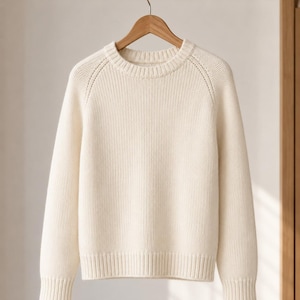 Könnte beinhalten: Ein cremefarbener Langarmpullover mit geripptem Rundhalsausschnitt, Bündchen und Saum. Das Strickkleidungsstück hat Raglanärmel mit einem dezenten vertikalen Stichmuster entlang der Schulternähte und hängt an einem Holzkleiderbügel.