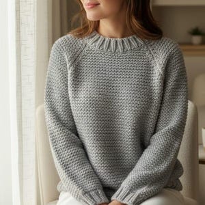 May include: A light grey long-sleeve jumper featuring a textured knit pattern, a ribbed crew neckline, and ribbed cuffs. The garment has raglan sleeves and a consistent knit structure throughout the body, providing a simple and uniform appearance.