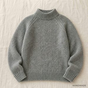 May include: A grey long-sleeve jumper featuring a mock neck collar, ribbed cuffs, and a ribbed hem. The knit garment is constructed with raglan sleeves and displays a consistent textured stitch pattern throughout the entire piece.