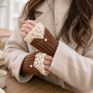 May include: A pair of brown ribbed fingerless gloves featuring a cream-colored crocheted lace trim at the cuffs and a single small white pearl accent centered on the top of each glove, providing a decorative detail to the knit fabric construction.