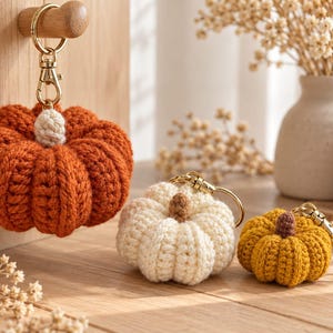 May include: Three crocheted pumpkin keychains in orange, cream, and mustard yellow. Each pumpkin features a small stem in a contrasting color and is attached to a gold-toned metal keyring with a lobster clasp for secure attachment to items.
